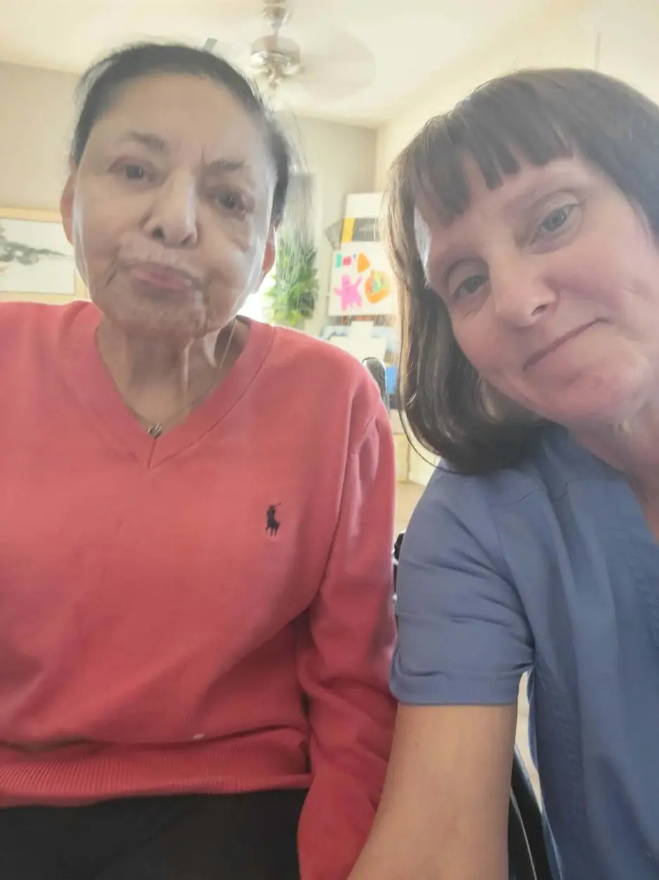 A caregiver in blue scrubs sitting with an elderly client in a home living room